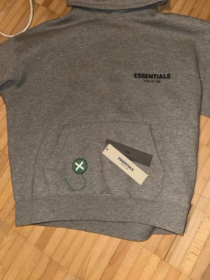 Grå hoodie från Essentials Fear of God - Snygg grå hoodie från Essentials Fear of God med svart logga på bröstet. Klassisk känguruficka framtill och ribbade muddar. Tillverkad i mjukt bomullsmaterial och har en avslappnad passform. Perfekt för en streetwear-look.