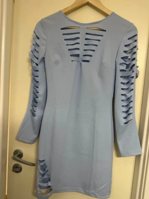 Ljusblå klänning med cut out-detaljer - Trendig ljusblå klänning med långa ärmar och coola cut out-detaljer längs ärmar, bröst och höft. Figurnära passform och kort längd, perfekt för dig som vill sticka ut. Tillverkad i mjukt material som känns skönt mot huden. Onesize (passar nog xs-m