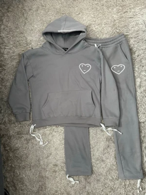 Grått mjukisset från carsicko - Säljer ett grått mjukisset med hoodie och byxor från carsicko. Hoodien har en vit hjärtdetalj med smiley på bröstet, känguruficka och dragsko i nederkant. Byxorna har resår i midjan och matchande hjärtdetalj. Setet är i mjukt bomullsliknande material.