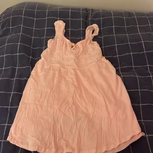 Ljusrosa kort klänning med volang - Söt ljusrosa kort klänning med breda axelband och volangdetaljer. Klänningen har en luftig passform och är tillverkad i ett tunt, skrynklat tyg som ger en avslappnad känsla. Perfekt för varma dagar och sommarens alla tillfällen.