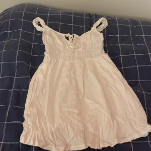 Vit kort klänning med snörning - Söt vit kortklänning med snörning framtill och breda axelband. Klänningen har en lätt utställd kjol och är tillverkad i ett tunt, mjukt tyg som ger en luftig känsla. Perfekt för varma dagar och enkel att styla med accessoarer.