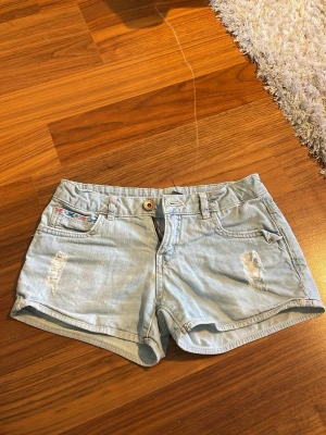 Ljusblå jeansshorts från Zara  - Säljer ett par ljusblå jeansshorts från Zara Kids med slitna detaljer och med broderade detaljer.                                                                     Skulle säga att de är lite större än 140. Midjemåttet: 35 cm (mätt platt)                          Höjden över rumpan: 27 cm (mätt platt) 