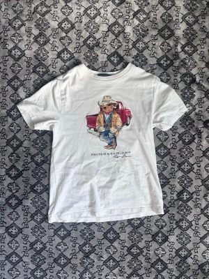 Ralph lauren t-shirt - Denna t-shirt från Ralph lauren med en nallebjörn som ett tryck på framsidan. är i ganska bra skick. Det finns en fläck som inte riktigt går bort i vanlig tvättmaskin men om man ger det något extra så borde det gå super. Den ska passa ungdomar mellan 13-16. Men skulle säga att den sitter som S.
