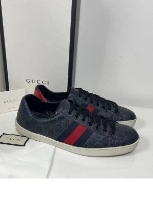 Gucci Ace sneakers med GG-mönster - Snygga Gucci Ace sneakers i mörkblått läder med GG Supreme-mönster, klassiska ränder i rött och marinblått på sidan samt kontrasterande hälflikar i rött och blått ormskinn. Vita sulor och svarta skosnören ger en clean look. Perfekta för dig som gillar lyxiga detaljer.