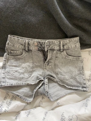 Grå lågmidjade denimshorts med broderade fickor - Snygga grå denimshorts med låg midja och dubbla knappar framtill. Shortsen har fram- och bakfickor, där bakfickorna är dekorerade med ett broderat hjärtmönster. Perfekta för sommardagar och har en klassisk jeanslook.