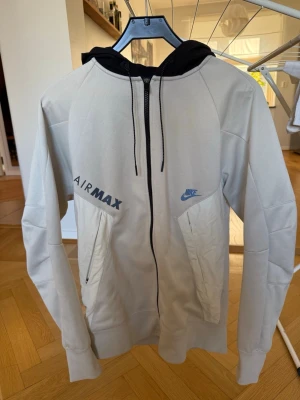 Nike Air Max beige hoodie med dragkedja - Snygg beige hoodie från Nike Air Max med svart huva och dragkedja framtill. Två stora fickor med dragkedja och tryckta loggor på bröstet. Perfekt för en sportig och avslappnad stil. Materialet känns mjukt och bekvämt. Passar bra till S/M