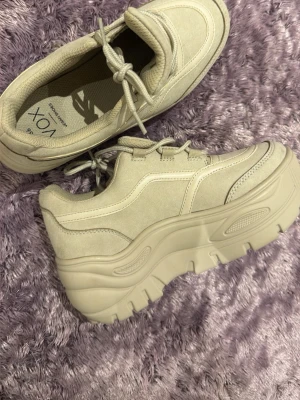 Beige chunky sneakers från VOX - Säljer ett par beige chunky sneakers från VOX med kraftig platåsula och snörning. Skorna har en mix av mockaimitation och textil, samt detaljerade sömmar för en trendig look. Perfekta för dig som gillar streetstyle och vill ha extra höjd.