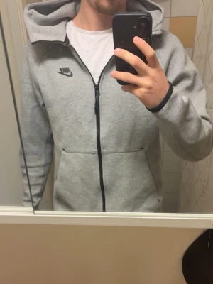 Nike teck fleece - Säljer ett par gråa mjukisbyxor med dragsko i midjan och muddar vid bensluten. Byxorna har en avslappnad passform och är tillverkade i mjukt sweatshirtmaterial, perfekta för chill dagar. En liten logga syns på ena benet.