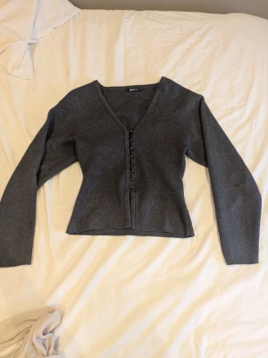 Mörkgrå kofta från Gina Tricot - Snygg mörkgrå kofta från Gina Tricot med v-ringning och knappar framtill i storlek XS. Modellen är figurnära med långa ärmar och mjukt, stickat material. Perfekt att matcha med jeans eller kjol för en clean look.