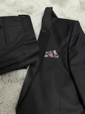 Svart kostym med kavaj och byxor - Säljer en klassisk svart kostym med matchande kavaj och byxor. Kavajen har två knappar, spetsiga slag och en snygg bröstficka med färgglad näsduk. Materialet känns slätt och elegant, perfekt för dig som vill ha en stilren look.