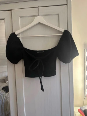 Svart croppad topp från Zara - Trendig svart croppad topp från Zara med puffiga korta ärmar och snygg knytning framtill. Toppen har en ribbad struktur och offshoulder-design som ger en modern vibe. Perfekt att styla med höga jeans eller kjol.