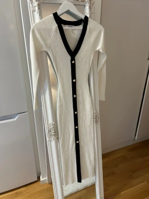 Vit långklänning med svarta detaljer - Säljer en ribbad vit långklänning med svarta kanter och knappar framtill. Klänningen har lång ärm, v-ringning och är figurnära i modellen. Perfekt för dig som gillar stilrena kontraster och enkel elegans.