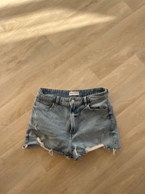 Blå denimshorts från Zara - Säljer ett par klassiska blå denimshorts från Zara med råa, fransiga kanter och slitningar framtill. Shortsen har fem fickor, knappgylf och normal passform. Perfekta för varma dagar och en avslappnad stil.