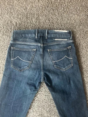 Jacob cohen jeans - Säljer ett par blå slim fit jeans med snygga kontrastsömmar och klassiska bakfickor. Jeansen har bälteshällor och en något tvättad look som ger en avslappnad vibe. Perfekta för dig som gillar en smalare passform och stilren design. Väldigt bra skick, bara lappen där det brukar stå Jacob cohen är sönder, men inget man tänker på