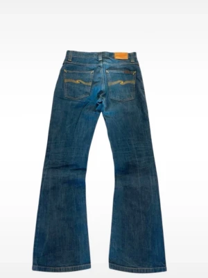 bootcut jeans Nudie Jeans - Säljer ett par klassiska blå bootcut jeans från Nudie Jeans med snygga kontrastsömmar och broderade bakfickor. Jeansen har normal midja och är tillverkade i slitstark denim. Perfekta för en avslappnad och trendig look.