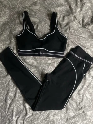 Svart Alo set med vita detaljer - Snyggt svart set från Alo med sport-bh och tights. Setet har vita kontrastsömmar och logga framtill på både bh och byxa. Materialet är stretchigt och följsamt, perfekt för både träning och chill. Modern design med breda axelband och hög midja.
