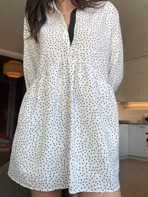 Vit prickig skjortklänning - Vit kort skjortklänning med svarta prickar och knappar framtill. Långärmad modell med avslappnad passform och lätt rynkad midja. Perfekt för dig som gillar en enkel men trendig look.
