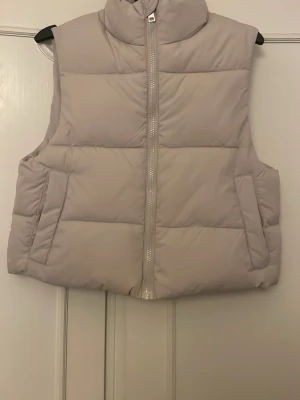 Ljusbeige dunväst från 157 XS - Snygg ljusbeige dunväst från 157 i storlek XS. Västen har hög krage, dragkedja framtill och två fickor på sidorna. Puffig modell som ger en trendig look och passar perfekt till lager-på-lager outfits. Tillverkad i ett mjukt och vadderat material.