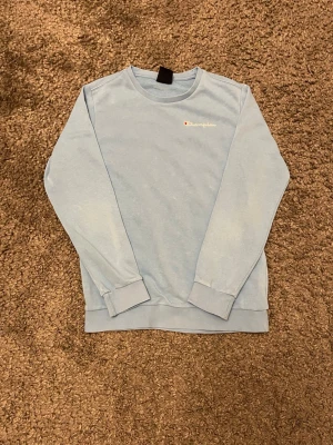 Ljusblå sweatshirt från Champion - Ljusblå sweatshirt från Champion med diskret logga broderad på bröstet. Tröjan har rund halsringning, långa ärmar och ribbade muddar vid ärmslut och nederkant. Perfekt för en avslappnad och sportig stil.