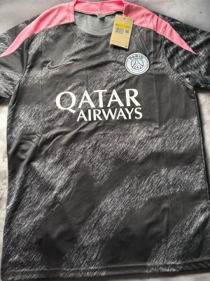 PSG matchtröja grå/rosa Nike - Snygg PSG matchtröja i grått med rosa axelpartier och vita detaljer. Tröjan har klubbmärke på bröstet, Qatar Airways-tryck och är tillverkad i ett lätt, ventilerande material. Perfekt för fotbollsträning eller att bära till vardags.