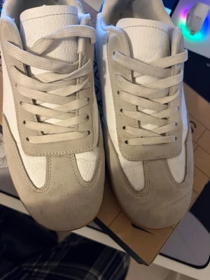 Masion margielas - Stilrena sneakers från maison mariella i beige mocka och vitt läder. Skorna har klassisk låg siluett, vita snören och en gummisula i brunt. Perfekta för dig som gillar en clean och tidlös look med en touch av retro.