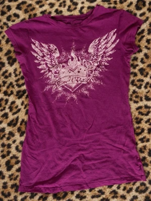 Y2k topp - Cool lila t-shirt med tryck av ett hjärta med eld och vingar på framsidan samt text på baksidan. T-shirten har korta ärmar och en figurnära passform. Perfekt för dig som gillar edgy och unika plagg.
