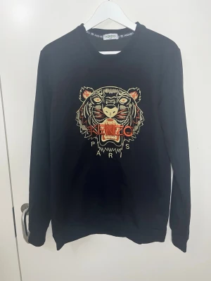 Svart sweatshirt med tigertryck Kenzo - Cool svart sweatshirt från Kenzo med det ikoniska tigerhuvudet broderat framtill i beige, orange och gult. Rund halsringning och långa ärmar. Perfekt statement-plagg för dig som gillar streetwear och vill sticka ut.