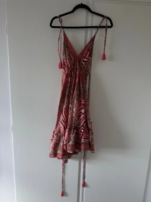 Mönstrad kortklänning - Säljer en super söt kortklänning med rosa och röda toner, zebramönster och tunna axelband med snörning och tofsar. Klänningen har omlott-look och volangkant nertill. Väldigt djup rygg. För referens: jag är 180cm (klänningen slutar strax över knäna)
