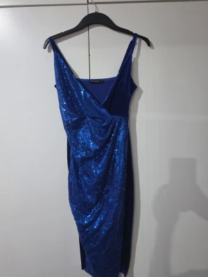 Blå glittrig omlottklänning - Snygg blå klänning med glittriga paljetter framtill och omlottdesign. Klänningen har smala axelband, v-ringning och slits framtill. Baksidan är i mjuk sammet. Perfekt för dig som vill sticka ut.