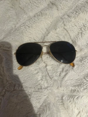 Guldiga aviator solglasögon - Snygga aviator solglasögon från Prestige med tunna guldfärgade metallbågar och mörka linser. Klassisk pilotform som ger en cool vibe och passar till de flesta outfits. Nässkydden är i transparent plast för extra komfort.