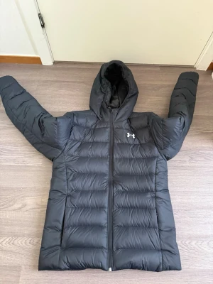 Svart dunjacka från Under Armour - Svart dunjacka från Under Armour med huva och quiltad design. Jackan har dragkedja framtill och logotyp på bröstet. Perfekt för kalla vinterdagar och har en sportig look som passar till många outfits.