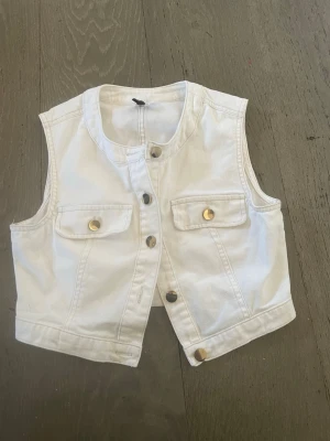 Vit jeansväst med guldknappar - Snygg vit jeansväst i croppad modell med rund halsringning och fyra guldknappar framtill. Västen har två bröstfickor med lock och guldfärgade knappar. Perfekt att styla över en t-shirt eller klänning för en trendig look.