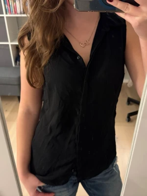 Svart ärmlös blus med krage - Säljer en svart ärmlös blus med klassisk krage och knäppning framtill. Perfekt till jeans eller kjol för en clean och enkel look. Materialet känns lätt och svalt, vilket gör den skön att bära under varmare dagar.