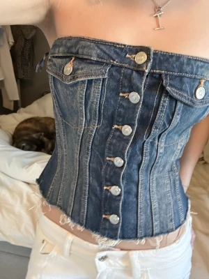 Mörkblå denim korsett topp - Trendig mörkblå korsett topp i denim med silverfärgade knappar framtill och råa kanter nedtill. Toppen är axelbandslös och har dekorativa sömmar samt fickdetaljer upptill. Perfekt för en edgy och cool look.