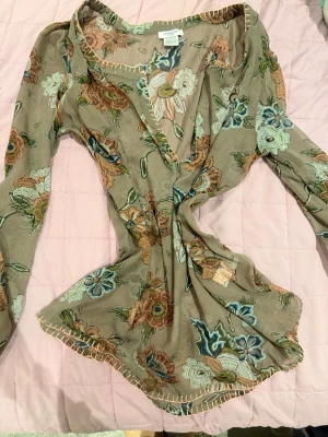 Blommig omlottblus från Topshop - Säljer en beige omlottblus från Topshop med blommigt mönster i grönt, blått och brunt. Blusen har djup v-ringning, långa ärmar och dekorativa sömmar längs kanterna. Perfekt för en trendig och avslappnad look.