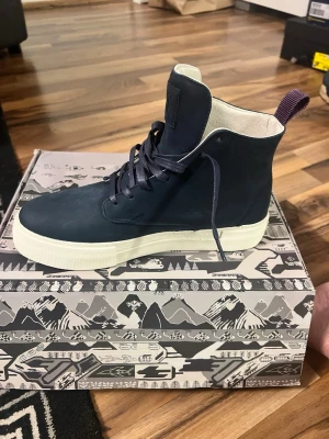 Mörkblå höga sneakers från Eytys - Snygga mörkblå höga sneakers från Eytys med vit tjock sula och lila hälflik. Skorna har en minimalistisk design i slätt läder och klassisk snörning. Eytys-logga bak på hälen. Perfekta för dig som gillar streetwear och stilrena detaljer.