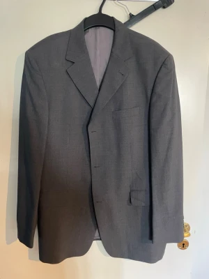 Klassisk grå kavaj med enkelknäppning - Stilren grå kavaj med enkelknäppning och klassisk krage. Kavajen har två framfickor med lock och en bröstficka. Fodrad insida och rak passform ger en tidlös look. Perfekt till skjorta och jeans eller mer uppklätt. Materialet känns som ullblandning.