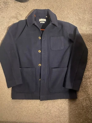Mörkblå overshirt från Strömberg  - Stilren mörkblå overshirt från Strömberg med klassisk krage, tre utanpåliggande fickor och knäppning framtill. Tillverkad i mjuk ull. Perfekt lager-på-lager-plagg för en avslappnad men snygg look.
