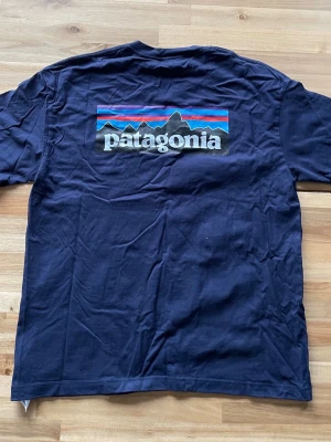 Mörkblå Patagonia t-shirt, storlek L - Snygg mörkblå t-shirt från Patagonia med klassisk logga och bergmotiv i färg på ryggen samt liten logga på bröstet. Tillverkad i mjuk bomull och har en normal passform med korta ärmar.