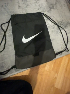 Svart gympåse från Nike - Snygg svart gympåse från Nike med klassisk vit logga på framsidan. Väskan har dragskostängning och tunna axelband samt en liten ficka med dragkedja. Perfekt för träning eller skola, enkel att bära och rymmer det viktigaste.