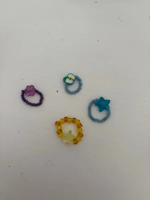Färgglada pärlringar med blommor och stjärnor - Fyra handgjorda ringar av små pärlor i olika färger: lila, blå, gul och turkos. Varje ring har en dekoration i form av en blomma eller stjärna. Perfekta för att addera en lekfull touch till din stil.