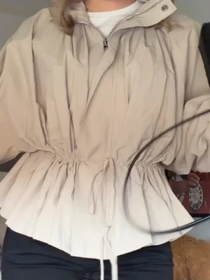 Beige jacka med snörning i midjan - Jättetendig slutsåld och snygg beige vindjacka med dragkedja och hög krage. Jackan har snörning i midjan för en justerbar passform och långa ärmar. Perfekt för dig som vill ha en stilren och funktionell jacka till vardags. Slutsåld på hela hemsidan