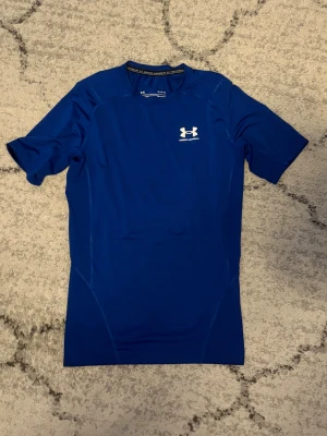 Blå träningströja från Under Armour - Snygg blå funktions-tshirt från Under Armour med korta ärmar och logga på bröstet. Tillverkad i ett stretchigt och snabbtorkande material som passar perfekt till träning och sportaktiviteter. Tight passform för en sportig look.