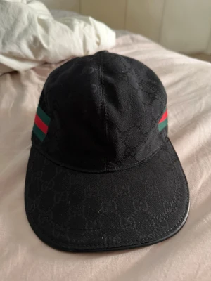 Svart Gucci keps med GG-mönster - Snygg svart keps från Gucci med klassiskt GG-monogram i tyget och de ikoniska gröna och röda ränderna på sidorna. Justerbar rem bak med gulddetalj. Perfekt accessoar för dig som vill ha en exklusiv och trendig look.