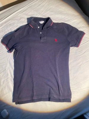 Mörkblå piké från Ralph Lauren - Klassisk mörkblå pikétröja från Ralph Lauren med röd logga på bröstet. Kragen har vita och röda detaljer och tröjan har korta ärmar. Tillverkad i mjuk bomull som känns skön mot huden. Endast en liten fläck på bröstet annars bra skick