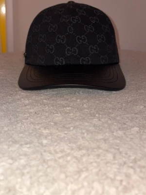 Svart keps från Gucci med GG-mönster - Snygg svart keps från Gucci med klassiskt GG-monogram i grått över hela tyget. Skärmen är i svart läder-look och kepsen har en stilren och modern form. Perfekt accessoar för dig som gillar exklusiva märken och streetwear.