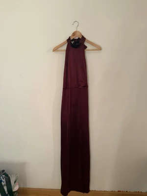 Vinröd långklänning med öppen rygg - Säljer en  vinröd långklänning i satin material. Klänningen har hög halsringning och öppen i rygg. Perfekt balklänning eller för festliga tillfällen.