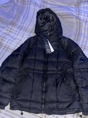 Svart dunjacka från C.P. Company - Snygg svart dunjacka från C.P. Company med huva och dragkedja framtill. Jackan har två sidofickor med dragkedja och den ikoniska linsen på ärmen. Perfekt för kalla dagar och ger en clean streetwear-look.