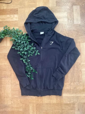 Svart zip-hoodie från Gymshark - Svart hoodie med dragkedja från Gymshark. Klassisk modell med huva och dragsko, samt fickor framtill. Gymshark-logga broderad på bröstet. Mjuk insida och ribbade muddar vid ärmslut och nederkant. Perfekt för en sportig och avslappnad stil.
