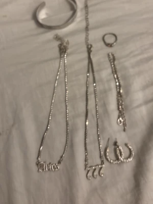 Smyckeset i silverfärg - Säljer ett set med accessoarer i silverfärg som inkluderar två halsband med textdetaljer, ett armband, en ring, ett stelt armband och ett par stora örhängen med gnistrande stenar. Perfekt för dig som gillar att mixa och matcha olika smycken.